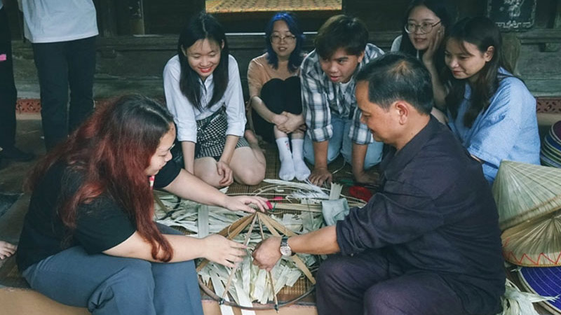 Nghệ nhân Lê Văn Tuy hướng dẫn nhóm bạn trẻ đan nón tại làng Chuông. Ảnh: QUÝ ĐỨC